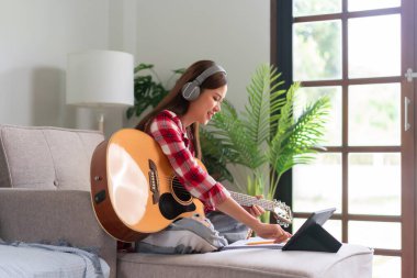 Hobi konsepti, genç Asyalı kadın kulaklık takıyor tablete müzik öğreniyor ve akustik gitar çalıyor..