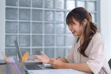 Günlük kıyafetli Asyalı genç bir öğrenci kulaklık takıyor ve bilgisayar başında öğretim görevlisi eğitimi alırken video konuşması yapıyor ve not defterine bilgi verileri yazıyor..