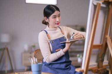 Genç Asyalı kadın sanatçı tuval üzerine resim çizmek için renk paleti kullanarak tüpten renk sıkıyor.