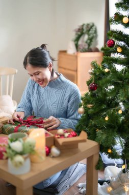 Kırmızı kurdele bağlamak için süveter giyen genç Asyalı kadın çam dalı ve böğürtlenlerle süslerken Noel bayramı ve kış mevsimleri için hediyeler hazırlıyor..
