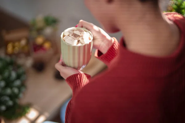 Young asian woman in sweater drinking hot chocolate with marshmallow while sitting near christmas tree in living room decorating ornaments for celebrate christmas festive holiday and winter seasons.