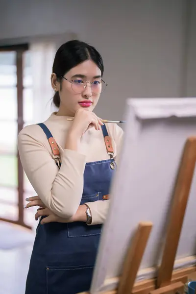 Young asian female artist hold paintbrush and looking on canvas to thinking create painting artwork.