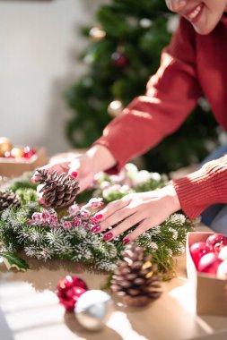 Ahşap masada Noel çelengi yapan genç Asyalı kadın ve çam kozalağı ve kırmızı böğürtlenlerle süslenirken Noel bayramı ve kış mevsimleri için süsleri hazırlayın..