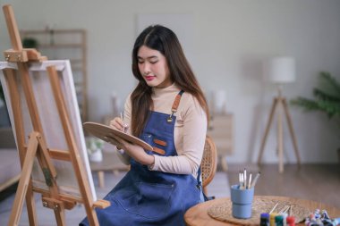 Genç Asyalı kadın sanatçı tuval üzerinde resim yapmak için renk paletiyle boya fırçası kullanıyor..