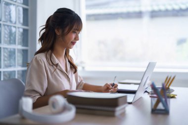 Gündelik giysiler içinde Asyalı genç bir öğrenci öğretim görevlileri eğitimini online olarak bilgisayar teknolojisiyle okurken aynı zamanda bilgi verileri yazıp üniversitede ödev yapıyor..