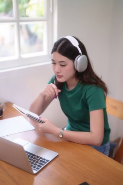 Genç Asyalı kadın öğrenciler kulaklık takarak çevrimiçi derste dizüstü bilgisayarda ağlar ve not defterlerine not alarak evden bilgi uzaktan eğitimini öğrenirler..