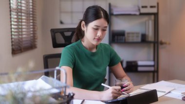 Genç Asyalı kadın öğrenciler ellerinde kahve bardağıyla dijital tabletle ders çalıştıktan sonra ev ödevlerini yazıyorlar ve internet üzerinden uzaktan eğitim alıyorlar..