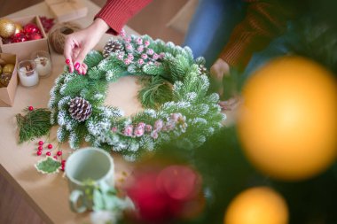 Noel çelengi ve kırmızı böğürtlenlerle süslenip Noel çelengi yapan Asyalı genç bir kadın Noel bayramı ve kış mevsimlerini kutlamak için süsleri evde hazırlıyor..