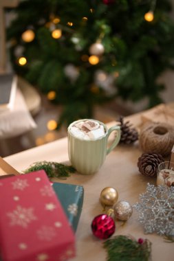 Noel ağacının yakınındaki ahşap masada marshmallow ve noel süsleriyle bir fincan sıcak çikolata Noel bayramını ve kış mevsimlerini kutlamak için hazırlanırken....