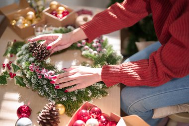 Ahşap masada Noel çelengi yapan genç Asyalı kadın ve çam kozalağı ve kırmızı böğürtlenlerle süslenirken Noel bayramı ve kış mevsimleri için süsleri hazırlayın..