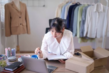 Genç Asyalı tüccar kadınlar çevrimiçi siparişleri ve müşteri bilgilerini bilgisayarda kontrol ediyorlar. İç işlerindeki çevrimiçi alışveriş işlerinde çalışırken ve nakliyat yaparken not almak için..