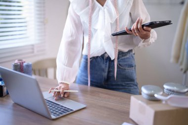 Genç Asyalı tüccar kadınlar online siparişleri ve müşteri bilgilerini kontrol etmek için dizüstü bilgisayarda veri yazarken tablete not alıyor ve merkez ofisteki çevrimiçi alışveriş işleri hakkında çalışıyor..