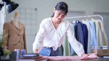 Genç Asyalı tüccar kadınlar, sandık boyundaki moda kıyafetlerinin ayrıntılarını kontrol etmek için ölçü bandı kullanırken tablete not alıyor ve merkez ofisteki çevrimiçi alışveriş işleriyle ilgili notlar alıyorlar..