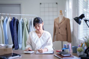 Genç Asyalı tüccar kadınlar hesap makinesi kullanarak çevrimiçi alışveriş işlerinin finansmanını hesaplıyor ve ev ofisteki müşterilere sevkiyat teslimatında çalışırken deftere bilgi yazıyorlar..