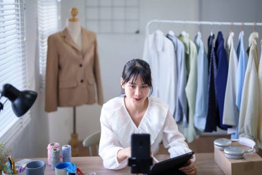 Genç Asyalı tüccar kadınlar video kaydetmek ve sosyal medyada kıyafet ürünleri satmak için akıllı telefon kullanırken dijital tablet satın alma siparişlerini kontrol ediyor ve çevrimiçi alışveriş işleri hakkında çalışıyorlar..