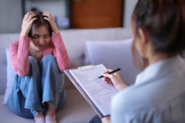 Asyalı hasta kadınlar endişe ve baş ağrısını vurgularken, kadın psikolog da psikolojik sağlık sorunlarını inceliyor ve akıl sağlığı terapisi hakkında danışmanlık için kağıda notlar yazıyor..