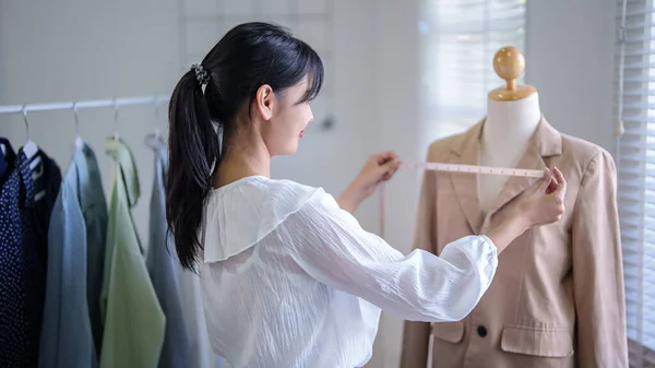 Young asian merchant women checking size details of new fashion clothes in mannequin with measuring tape while taking notes in clipboard and working about online shopping business in home office.