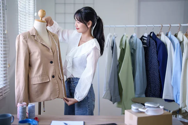 Young asian merchant women using measure tape to checking length size details of fashion clothes in mannequin while taking notes in clipboard and working about online shopping business in home office.