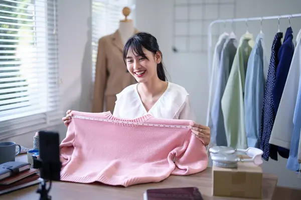 Young asian merchant women measuring size of clothes with measure tape to recording live streaming and sell clothes online in social media on smartphone while working about online shopping business.