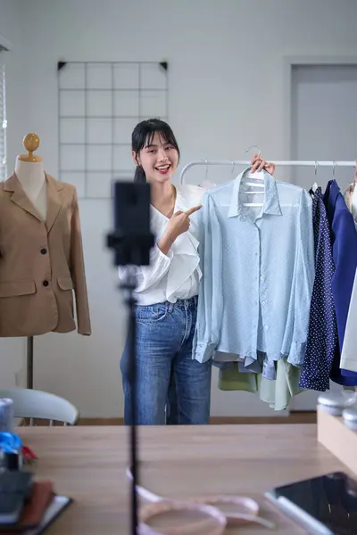 Young asian merchant women showing new fashion clothes to recording video and selling in social media on smartphone while working and shipping delivery for online shopping business in home office.