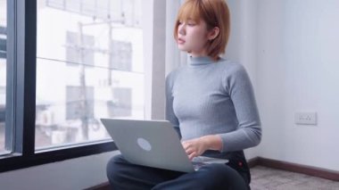 Ofisteki cam pencerenin yanında oturan ve özenle laptopuyla çalışan kendine güvenen Asyalı bir iş kadını odaklanmış ve kararlı bir tavır sergiliyor. Yüksek kalite 4k görüntü