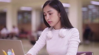Konsantre Asyalı genç kız öğrenci masada oturuyor ve online öğrenme dersi için dizüstü bilgisayar kullanıyor ve kütüphane kampüsünde sınava hazırlanırken notlar yazıyor.