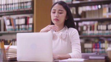 Konsantre Asyalı genç kız öğrenci masada oturuyor ve online öğrenme dersi için dizüstü bilgisayar kullanıyor ve kütüphane kampüsünde sınava hazırlanırken notlar yazıyor.