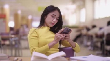 Asyalı güzel bir bayan öğrenci masada oturuyor ve cep telefonu kullanıyor ya da sosyal medyada mesajlaşıyor ya da kütüphanede ödev yaparken ya da kitap okurken şarkı dinliyor. Yüksek kalite 4k görüntü