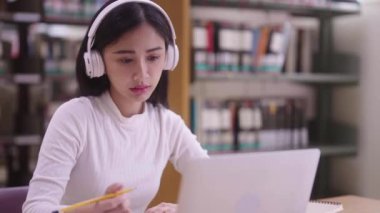Konsantre Asyalı genç kız öğrenci masada oturuyor ve online öğrenme dersi için dizüstü bilgisayar kullanıyor ve kütüphane kampüsünde sınava hazırlanırken notlar yazıyor.