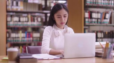 Konsantre Asyalı genç kız öğrenci masada oturuyor ve online öğrenme dersi için dizüstü bilgisayar kullanıyor ve kütüphane kampüsünde sınava hazırlanırken notlar yazıyor.