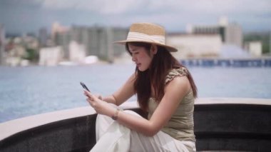 Asyalı bir kadın akıllı telefonunda sosyal ağ kullanıyor. Lüks yüzme havuzunun yanında rahatlayıp rahatlıyor. Tatil ve Teknoloji konseptinin her yerinde çalışıyor. Yüksek kalite 4k görüntü