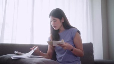 Genç Asyalı bir kadın hesap makinesi kullanarak kanepede oturur gelir ve giderleri hesaplar ya da aylık bütçeyi yönetir ve evdeki oturma odasındaki not defterine yazar. Yüksek kalite 4k görüntü