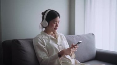 Genç Asyalı kadın, oturma odasında rahat bir kanepede otururken en sevdiği şarkıyı ya da müziği dinlerken ev hayatı konsepti olarak kulaklık takıyor. Yüksek kalite 4k görüntü