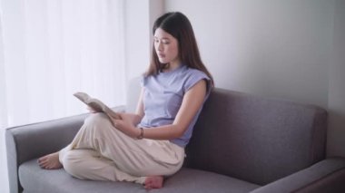 Çekici, genç Asyalı kadın rahat rahat kitap okuyor modern kanepenin önünde oturuyor oturma odasında dinleniyor ve rahatlıyor. Yüksek kalite 4k görüntü