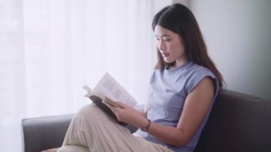 Çekici, genç Asyalı kadın rahat rahat kitap okuyor modern kanepenin önünde oturuyor oturma odasında dinleniyor ve rahatlıyor. Yüksek kalite 4k görüntü