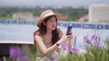 Asyalı bir kadın akıllı telefonunda sosyal ağ kullanıyor. Lüks yüzme havuzunun yanında rahatlayıp rahatlıyor. Tatil ve Teknoloji konseptinin her yerinde çalışıyor. Yüksek kalite 4k görüntü