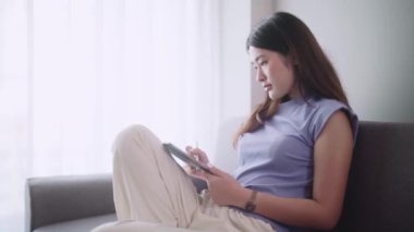Stylus kalem kullanan genç Asyalı kadın evdeki rahat koltukta otururken tablete proje verisi yazma fikri oturma odasındaki yaşam tarzı konsepti. Yüksek kalite 4k görüntü