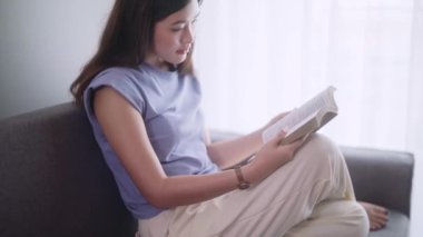 Çekici, genç Asyalı kadın rahat rahat kitap okuyor modern kanepenin önünde oturuyor oturma odasında dinleniyor ve rahatlıyor. Yüksek kalite 4k görüntü
