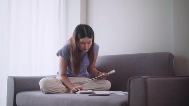 若いアジアの女性は 収入と費用を計算したり 毎月の予算を管理するために電卓を使ってソファーに座り 自宅のリビングルームのノートブックに書き込んでいます 高品質の4K映像 — ストック動画