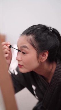 Asian woman applying mascara in front of a large mirror after bathing and styling her hair at home. Capture her beauty routine. High quality 4k footage