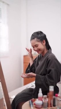 Asian woman applying face powder in front of a large mirror after washing and styling her hair at home. Showcase her beauty routine for enhanced sales and skincare awareness