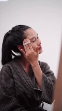 Asian woman applying face powder in front of a large mirror after washing and styling her hair at home. Showcase her beauty routine for enhanced sales and skincare awareness. High quality 4k footage