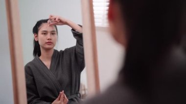 An Asian woman applying face cream to nourish her skin in front of a large mirror after washing and styling at home. Highlight the skincare routine. High quality 4k footage