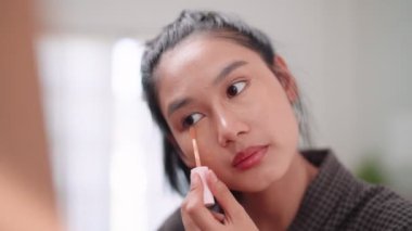Asian woman applying concealer on her face in front of a large mirror after washing and styling her hair at home. Capture her beauty routine. High quality 4k footage