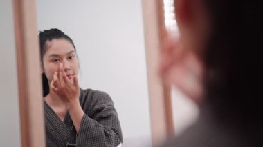 Asian woman applying concealer on her face in front of a large mirror after washing and styling her hair at home. Capture her beauty routine. High quality 4k footage