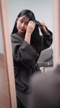 Asian woman using a hair straightener to achieve smooth hair in front of a large mirror after washing and styling at home. Showcase the beauty routine for enhanced sales. High quality 4k footage