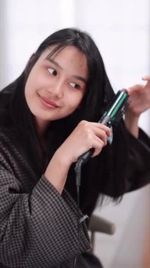 Asian woman using a hair straightener to achieve smooth hair in front of a large mirror after washing and styling at home. Showcase the beauty routine for enhanced sales. High quality 4k footage