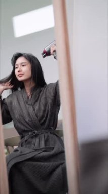 An Asian woman blow-drying her hair in front of a large mirror after washing and styling it at home. Capture the essence of beauty and self-care. High quality 4k footage