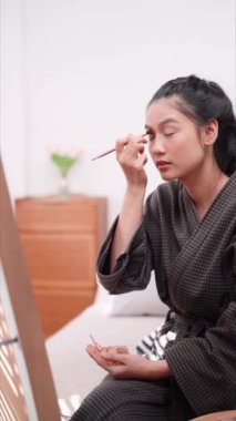 Asian woman applying eyeshadow in front of a large mirror after bathing and styling her hair at home. Showcase her beauty routine. High quality 4k footage