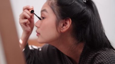 Asian woman applying mascara in front of a large mirror after bathing and styling her hair at home. Capture her beauty routine. High quality 4k footage